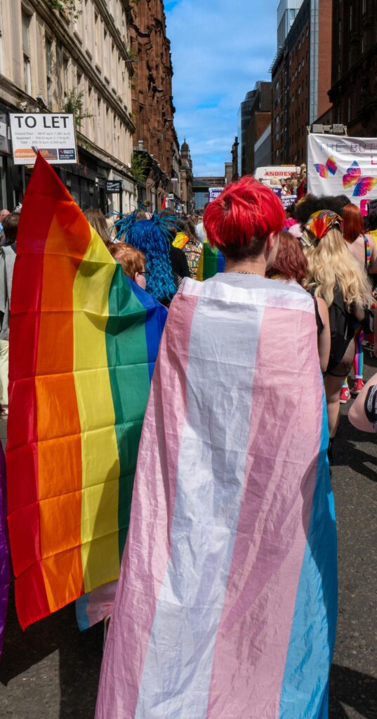 A person with red hair wearing the transgender flag on their back beside a person holding up a pride flag